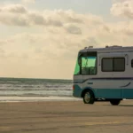 Wohnmobil am Strand, großes Reisemobil mit blauem unteren Bereich und Dekorlinie, Meereswellen im Hintergrund, Wolken am Himmel, sonniges Licht