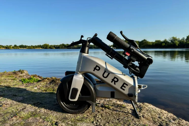 Der zusammengefaltete E-Scooter Pure Flex steht auf einem Felsen am Seeufer vor ruhigem Wasser.